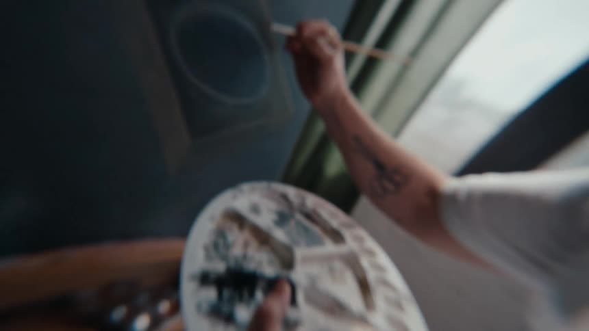 A close-up slow motion shot follows an artist's brush as it mixes paint on a palette and then applies it to a dark canvas