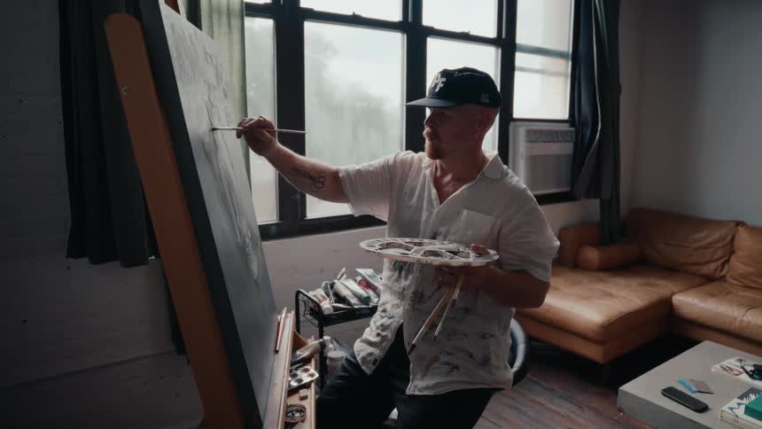 A focused male artist in a baseball cap paints on a large canvas in his naturally lit studio