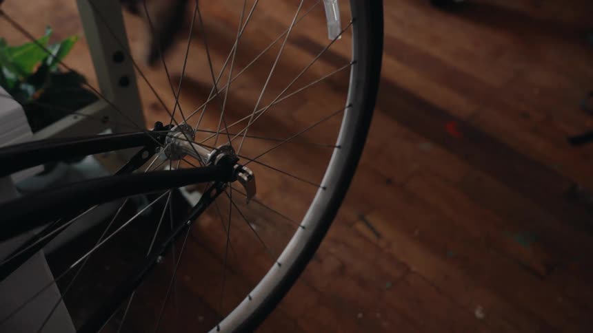 A close-up of a stationary bicycle wheel that starts to spin as a person walks by indoors on a wooden floor