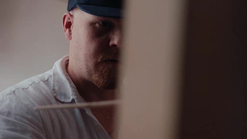 A focused artist in a cap and white shirt intently paints on a canvas in his studio, captured in a close-up shot