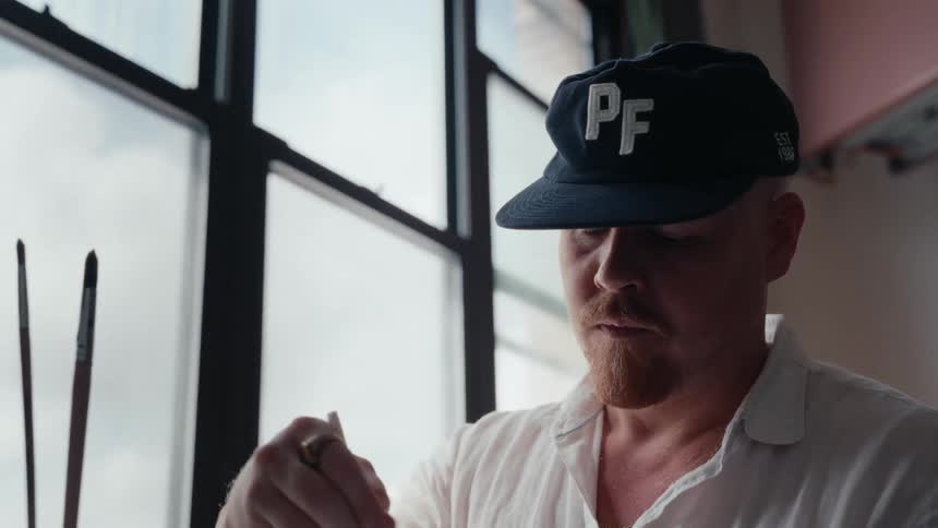 Close up of a focused male artist with a red beard intently painting in his naturally lit studio