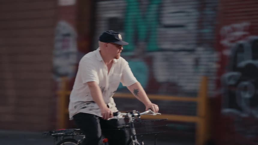 A stylish man with a palm tree tattoo rides his bicycle down a gritty urban street in slow motion