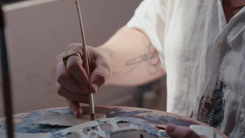A close-up shot of a tattooed artist's hand mixing paint colors on a wooden palette with a brush in a studio