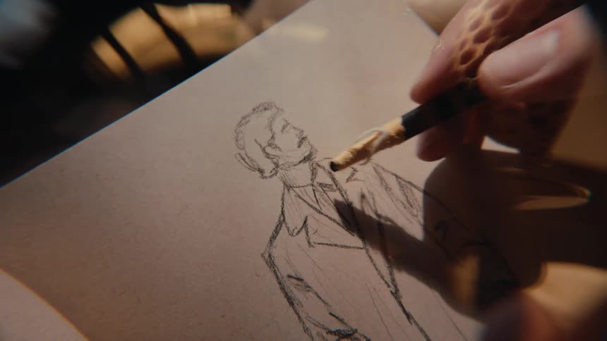 Close up of an artist's hand sketching a portrait of a man on paper with a pencil in warm, dappled light