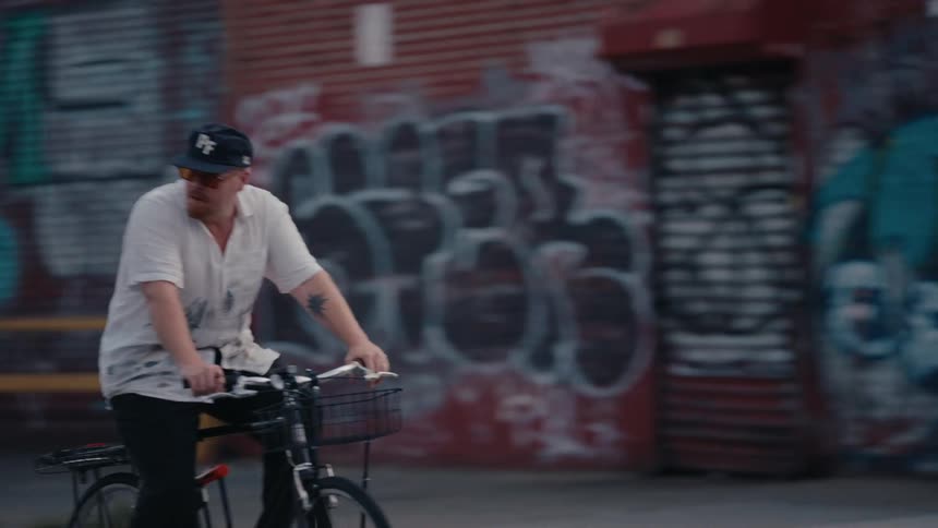 A man in a baseball cap rides his bicycle in slow motion down an urban street past graffiti-covered walls