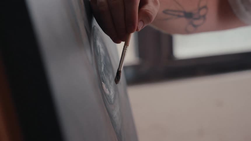 An artist with a tattooed arm carefully applies a brushstroke to an abstract painting in their studio