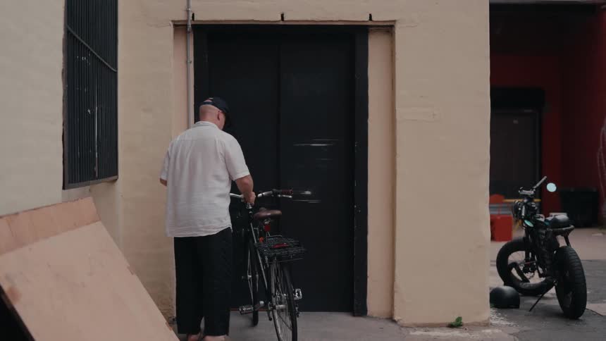 A man walks his bicycle up to an industrial freight elevator in an urban alleyway