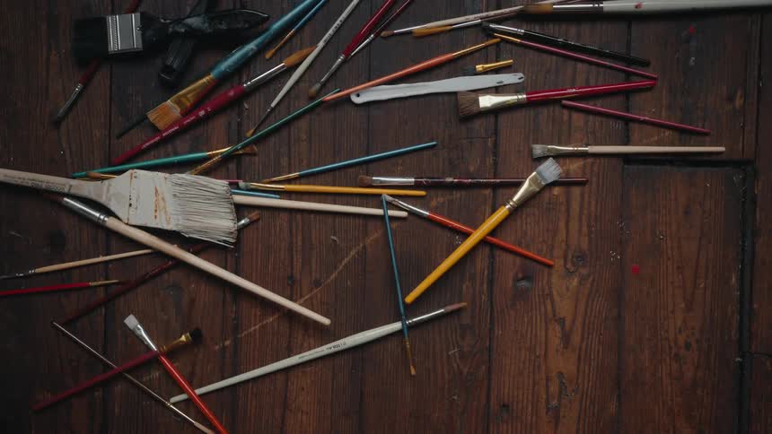 An assortment of colorful paintbrushes are dropped from above, scattering across a dark, rustic wooden floor