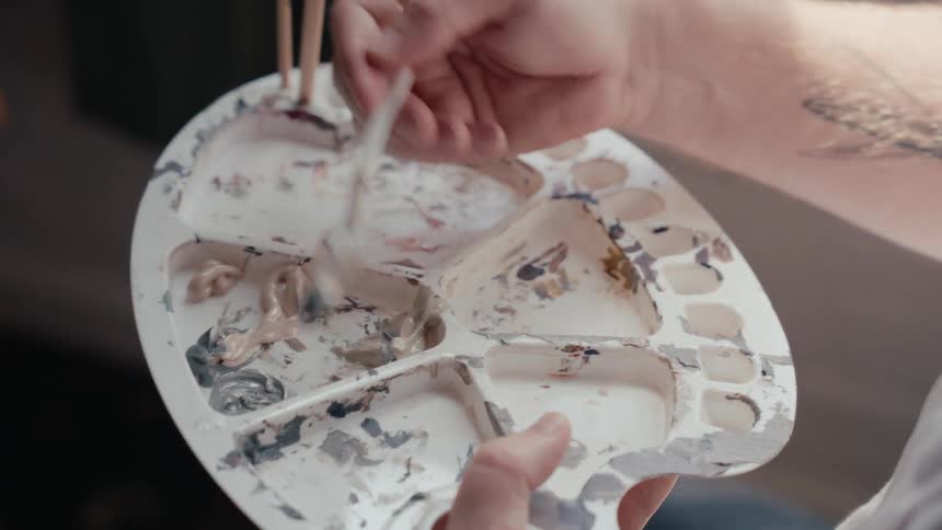 A close-up shot of an artist's hands with a tattoo mixing muted colors of paint on a well-used white palette