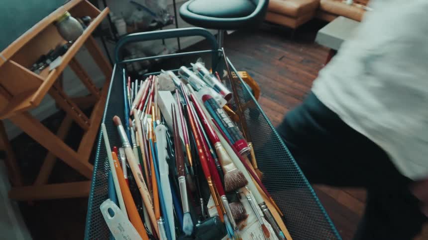 A collection of assorted paintbrushes sit in a mesh cart inside a rustic art studio as an artist prepares to paint