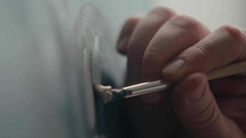 Extreme close-up shot of a painter's hand making a slow, precise brushstroke on a canvas