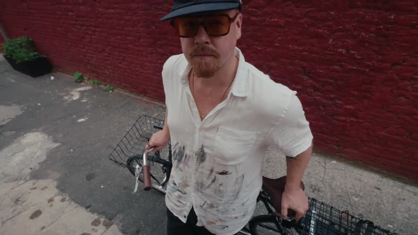 A stylish artist in a paint-splattered shirt and sunglasses stands with his bicycle in a city alley, looking up at the camera