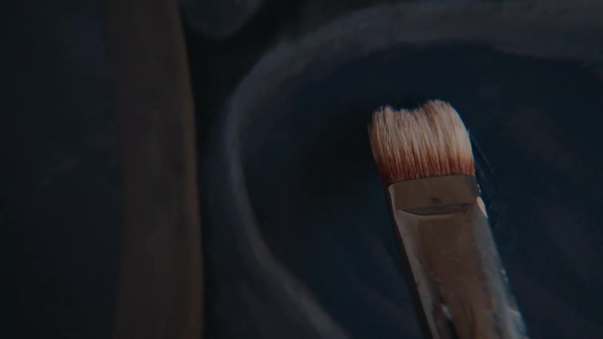 From the artist's perspective, a paintbrush is loaded with dark paint from a palette and applied to a canvas in a close-up shot
