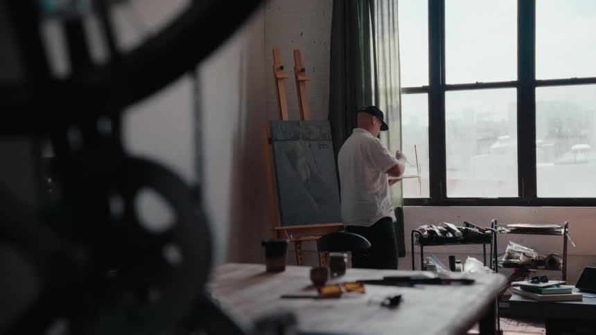 An artist walks over to his canvas on an easel and begins to paint in his naturally lit urban art studio