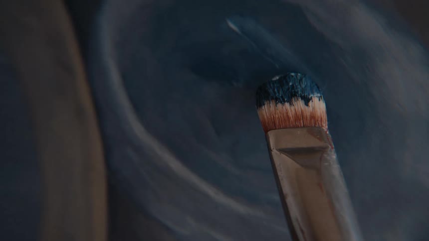 A macro, first-person view shows an artist dipping a brush into a paint palette and then applying dark blue paint to a canvas in broad strokes