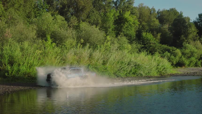 A light blue pickup truck drives through a river in slow motion, splashing water along the tree-lined bank