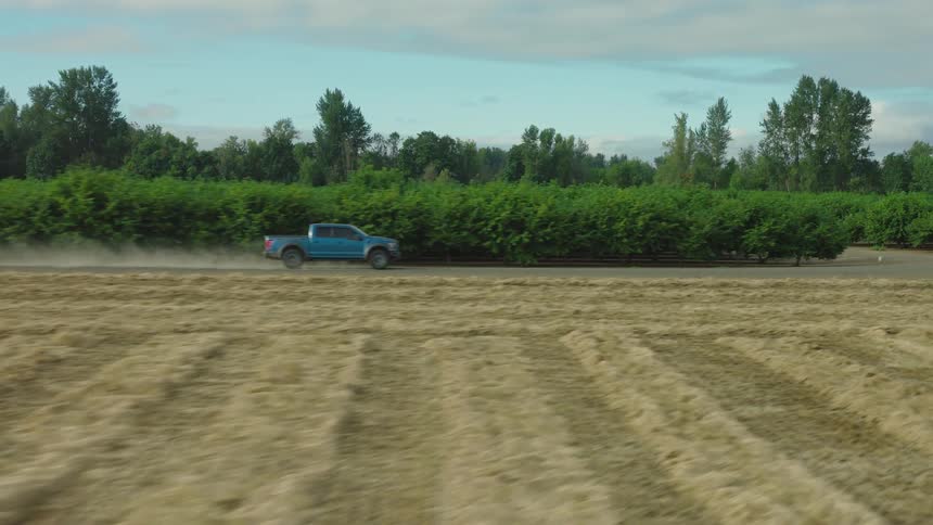 An aerial shot tracks a blue pickup truck as it speeds down a dusty farm road next to a harvested field and a large orchard