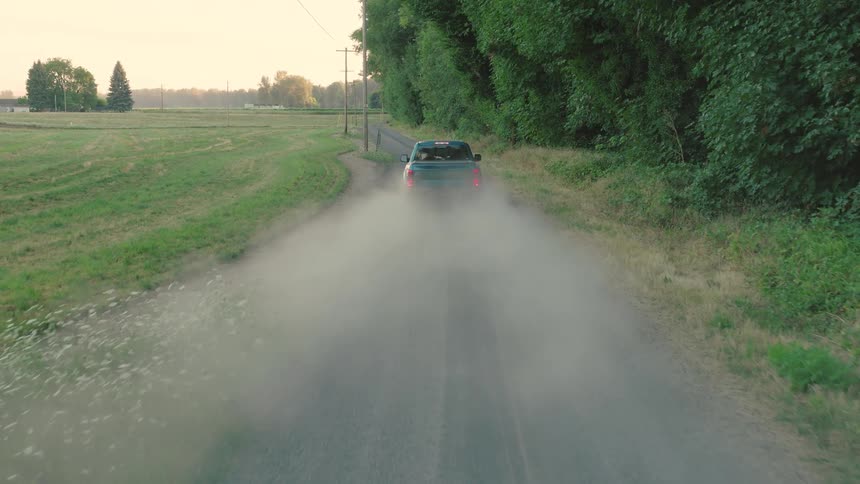 A blue Ford pickup truck kicks up a large cloud of dust as it drives away down a gravel country road at sunset