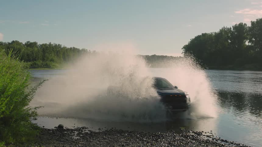 A powerful dark pickup truck splashes through a wide river surrounded by a lush forest in slow motion