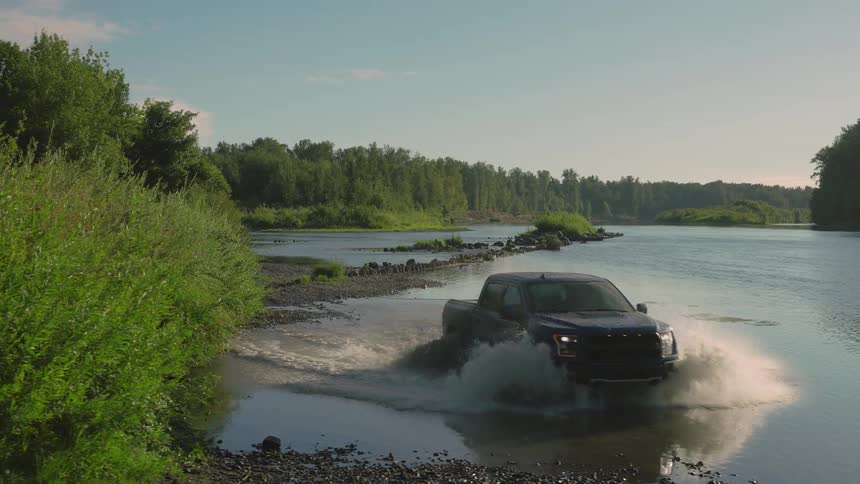 A black pickup truck splashes through a scenic river surrounded by lush green forest in slow motion