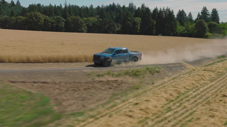 Aerial side-view of a blue pickup truck kicking up dust as it drives along a rural road next to golden fields on a sunny day