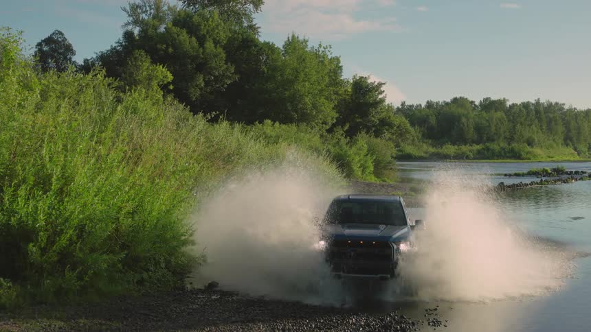 A powerful blue pickup truck drives through a river in slow motion, sending a huge spray of water into the air
