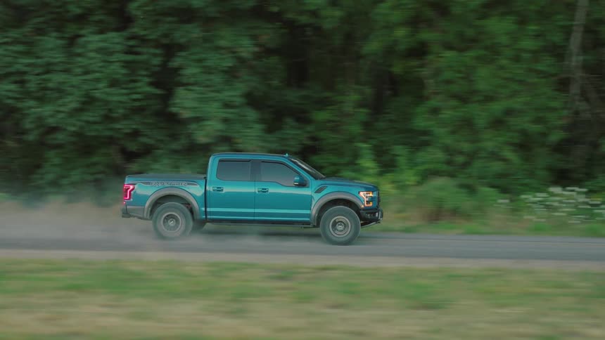 A teal pickup truck kicks up dust as it speeds down a rural dirt road with a backdrop of lush green trees