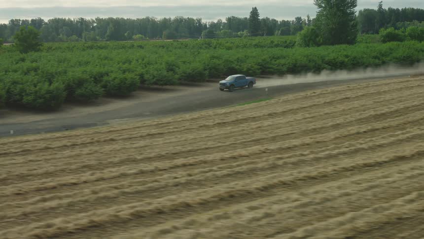 Aerial tracking shot of a blue pickup truck speeding down a dusty dirt road next to sprawling farm fields