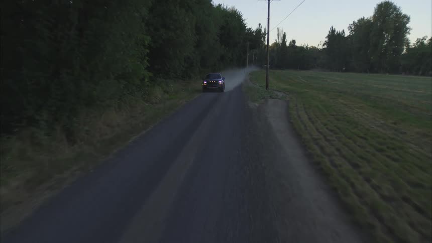 An aerial view shows a blue pickup truck driving down a dusty country road at dusk, kicking up dust as it passes