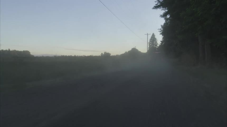 A powerful truck speeds down a gravel country road at dusk, kicking up a large cloud of dust in slow motion