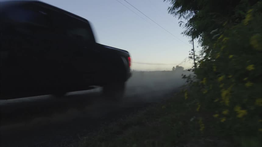 A pickup truck kicks up a large cloud of dust as it drives down a gravel road through the countryside at dusk