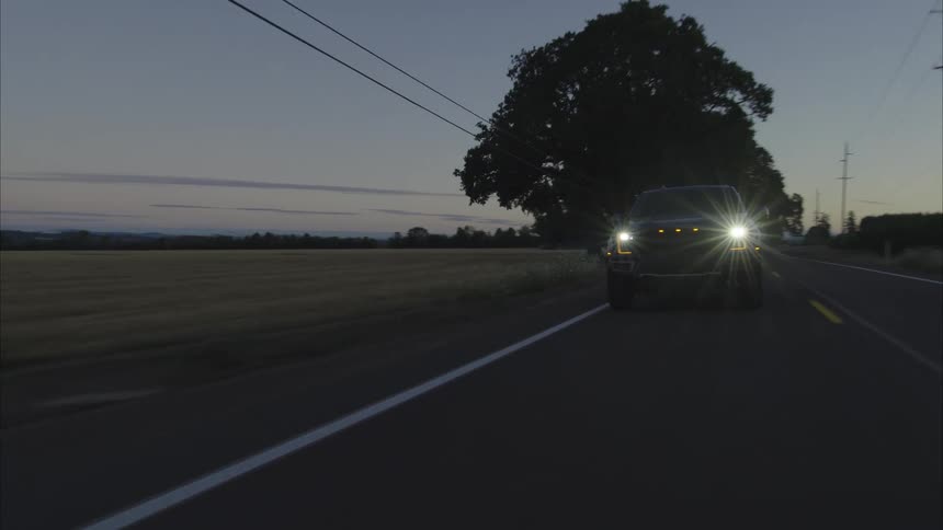 A dark pickup truck with bright headlights drives down a rural country road at dusk