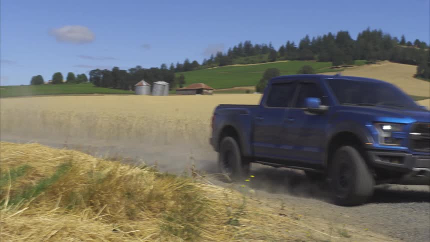 A blue Ford Raptor pickup truck kicks up dust as it drives in slow motion along a rural dirt road through rolling hills and wheat fields on a sunny day