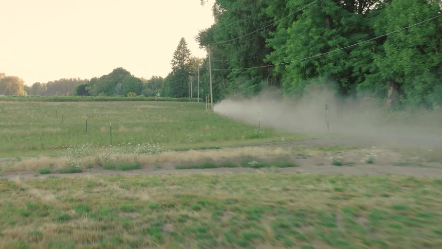 An aerial view tracks a car as it drives down a long, dusty rural road next to a green field at sunset