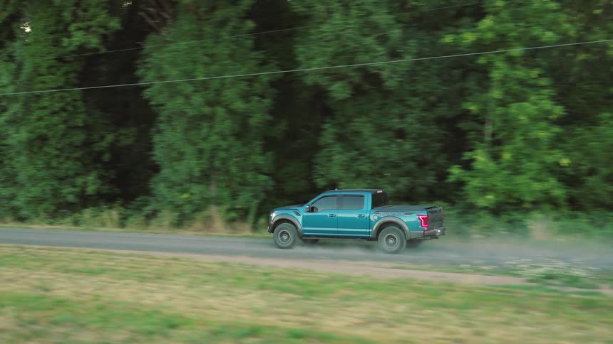 A blue Ford F-150 Raptor pickup truck drives down a rural country road kicking up dust with a lush green forest in the background