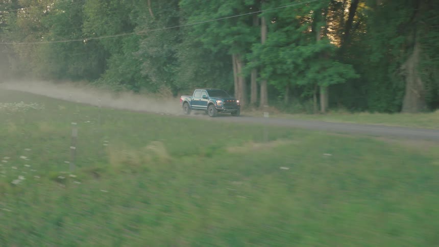 An aerial tracking shot follows a green pickup truck driving down a dusty country road next to a farm at sunset
