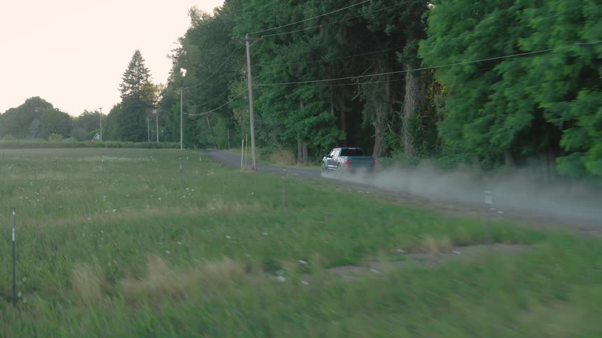 A blue pickup truck kicks up dust as it drives down a rural country road next to a field and forest during a hazy sunset