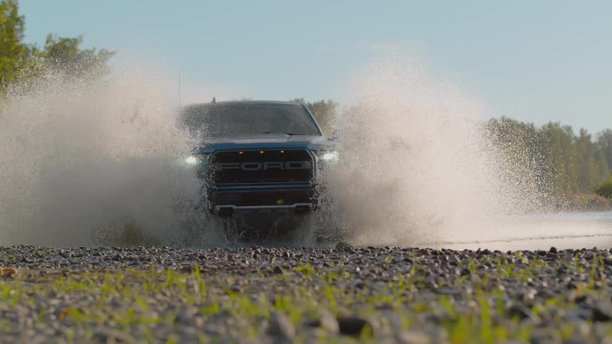 A powerful blue Ford truck drives head-on through a river, creating a huge splash in dramatic slow motion