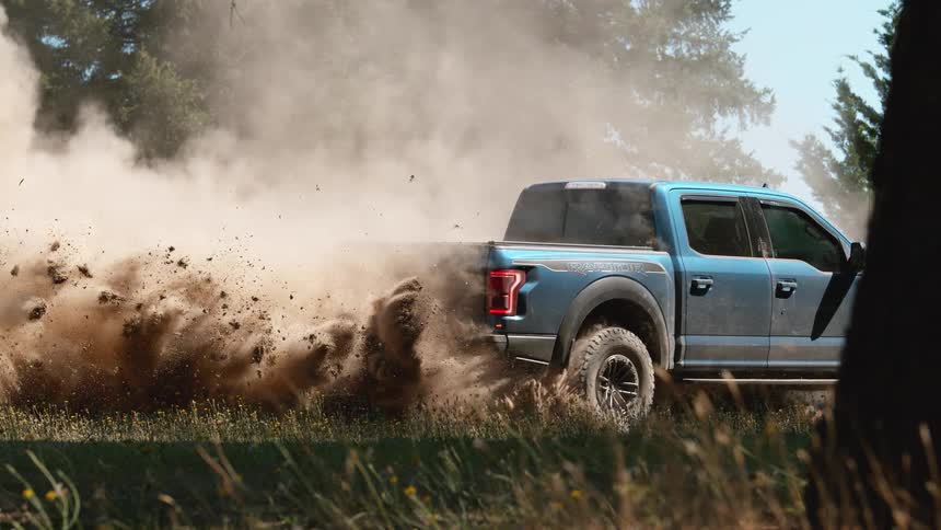 A blue off-road truck spins its wheels in a field, kicking up a massive cloud of dirt in super slow motion
