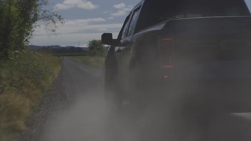 A dark blue pickup truck kicks up a cloud of dust as it drives down a gravel country road in slow motion