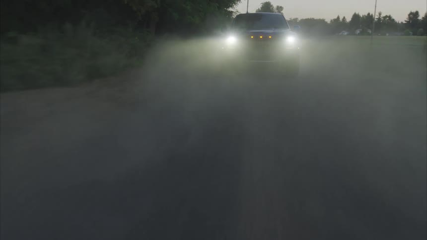 Aerial drone follows a powerful Ford Raptor truck with its headlights on as it drives down a dusty country road at dusk