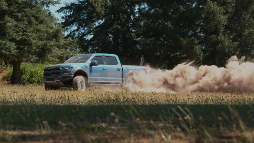 A blue off-road truck spins its tires in slow motion, creating a large cloud of dust in a grassy field