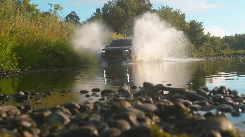 Slow motion, low-angle shot of a powerful black pickup truck driving through a river, creating a dramatic splash