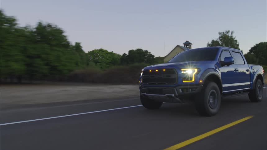 A powerful blue Ford F-150 Raptor drives along a winding rural road at dusk with its headlights brightly lit