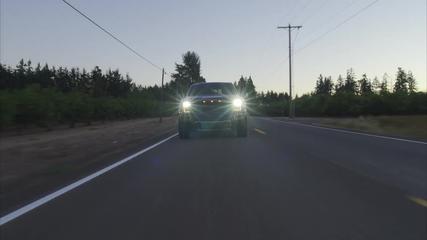 A powerful pickup truck with its headlights on drives down a quiet country road at dawn, transitioning from darkness into the early morning light