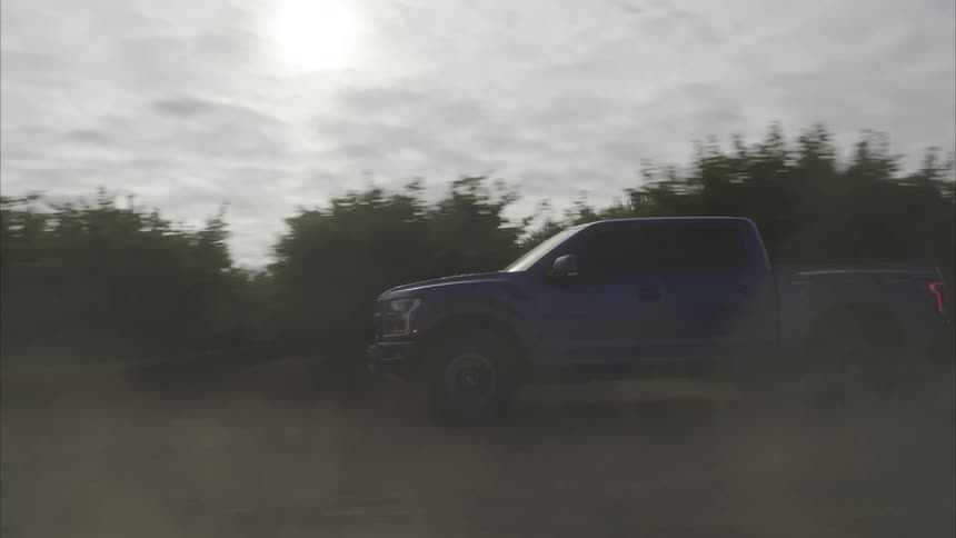 A blue Ford F-150 Raptor pickup truck kicks up dust while driving down a dirt road in the countryside