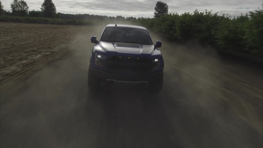 A dramatic front-facing slow-motion tracking shot shows a blue pickup truck driving fast down a dusty dirt road in the countryside