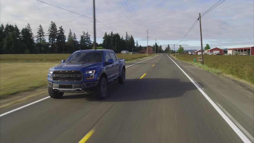 A blue Ford Raptor pickup truck drives along a rural road, passing farms and a classic red barn under a partly cloudy sky