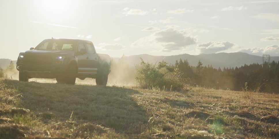 A powerful blue pickup truck drives off-road through a dusty, sunlit mountain field in slow motion