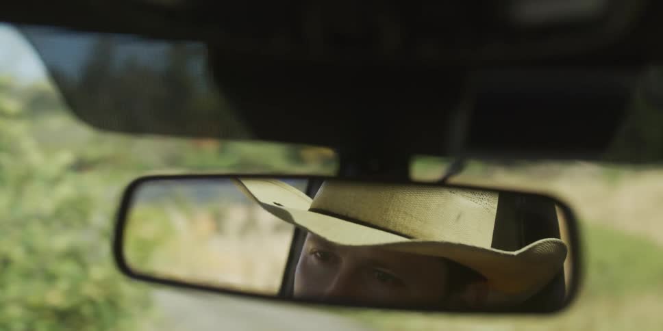 A man wearing a cowboy hat is reflected in the rearview mirror of a car as he drives along a rural country road in slow motion